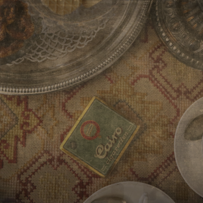 Lähikuva kuviollisesta pöytäliinasta, jonka päällä "Cairo"-savukerasia. Kuvan reunoilla posliinisia ja hopeisia astioita.