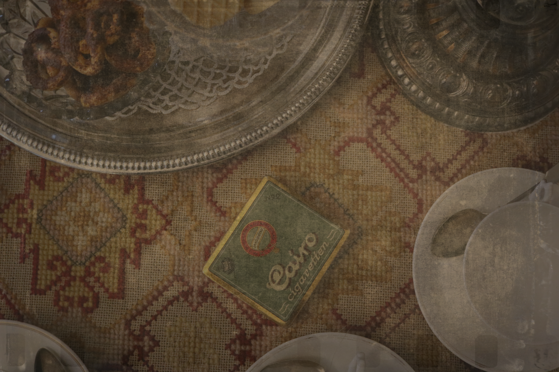 Lähikuva kuviollisesta pöytäliinasta, jonka päällä "Cairo"-savukerasia. Kuvan reunoilla posliinisia ja hopeisia astioita.